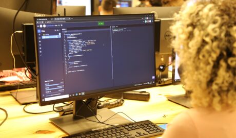 Aluna com cabelos cacheados claros concentrada programando em um computador. A tela exibe código, indicando a formação em desenvolvimento de software.