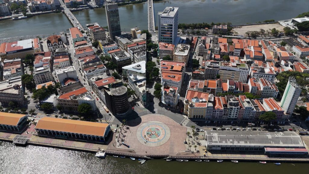 Vista aérea diurna do Bairro do Recife, com edifícios históricos de telhados alaranjados contrastando com estruturas mais modernas, e o rio que o contorna, simbolizando a revitalização e o crescimento do Porto Digital