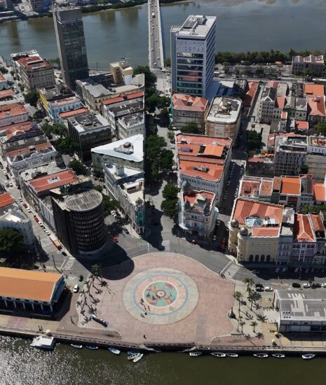 Vista aérea diurna do Bairro do Recife, com edifícios históricos de telhados alaranjados contrastando com estruturas mais modernas, e o rio que o contorna, simbolizando a revitalização e o crescimento do Porto Digital
